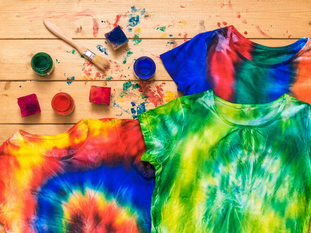 Three tie dye t-shirts on a wooden background with paint. Flat lay.